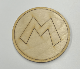 A wooden disk with a cut-out of the classic Mario letter "m" in the center, placed in the hole it was cut out of like a puzzle piece, with a ring cutout on the circumference of the circle, placed on a white surface.